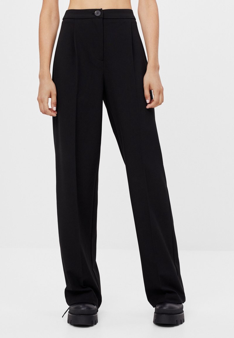 Pantalon noir taille haute à jambes larges avec plis sur le devant, fermeture par bouton, et chaussures compensées noires épaisses portés par une personne debout.