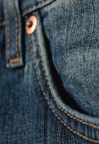 Close-up of blue denim pocket with visible copper rivet and orange stitching along pocket edge.