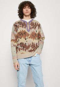 Suéter de punto con un patrón de camuflaje en marrón, beige y verde, con un acento morado y cierre de cremallera, y un corte holgado. Jeans de mezclilla azul claro.