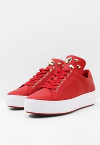 Baskets en cuir rouge avec une semelle plateforme blanche, des clous en forme de cœur dorés sur la languette, et des lacets plats rouges passant à travers des œillets dorés.