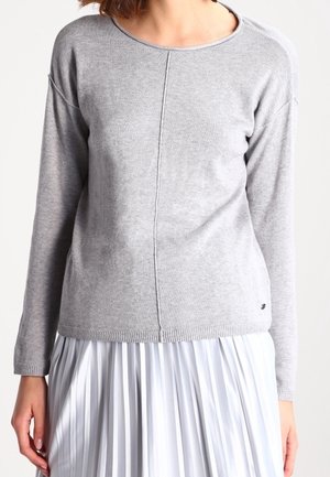 Kvinde iført en lysegrå langærmet sweater med en central søm, kombineret med et hvidt plisseret skørt.