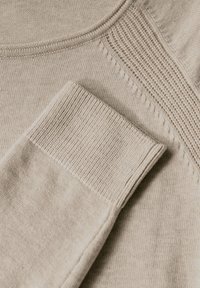 Beige gestrickter Pullover mit gerippten Bündchen und strukturierten Schultern. Glatter Stoff mit einem dezenten Muster und rundem Ausschnitt.