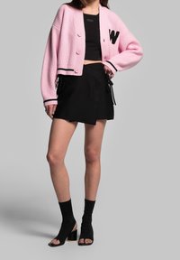 Růžový žebrovaný kardigan s černými pruhy a detaily v podobě písmen, doplněný o černou zavinovací sukni s pásky a černé sandály na podpatku s otevřenou špičkou.