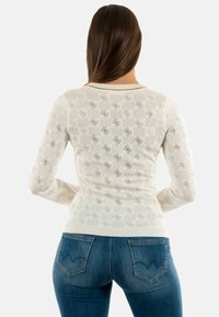 Top cu mânecă lungă de culoare ivorie, prezentând un model texturat și un design floral subtil, asociat cu blugi din denim albastru cu cusături distinctive pe buzunarele din spate.
