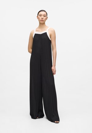 Woman standing wearing a sleeveless black pleated jumpsuit with white straps, feet partially visible in black open-toe shoes.