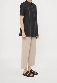 Persona che indossa una camicia nera a maniche corte con bottoni, pantaloni beige a gamba larga e sandali neri a doppia fascia, in piedi contro uno sfondo semplice.