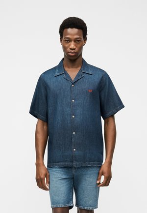 Ung mand iført mørk denimskjorte med korte ærmer, knapper og krave samt lyse denimshorts, stående foran en ensfarvet baggrund.