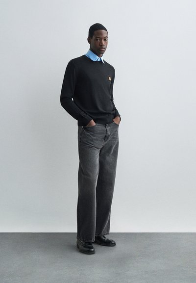 Mann står med hendene i lommene iført svart genser over blå skjorte, løse grå jeans og svarte dressko på grått gulv.