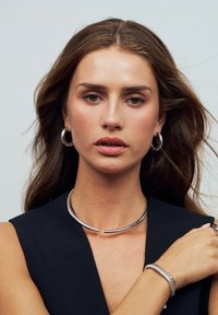 Sølv metal choker halskæde med et buet design. Model iført hoop øreringe og et sølv armbånd. Mørk ærmeløs top, neutral baggrund.