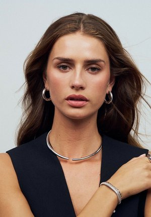 Silverfärgad metallchoker med en böjd design. Modell som bär hoopörhängen och ett silverarmband. Mörk ärmlös topp, neutral bakgrund.