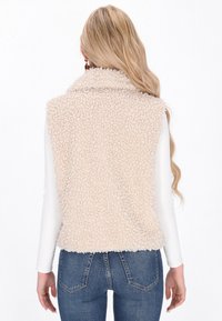 Mujer con cabello rubio largo de espaldas, vistiendo un chaleco sin mangas de felpa beige, una camiseta blanca de manga larga y jeans azules, con un fondo blanco.