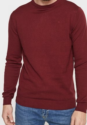 Homme portant un pull col rond à manches longues ajusté de couleur bordeaux, associé à un jean bleu, visible du cou aux hanches.