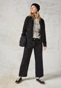 Giacca nera con cerniera e tasche frontali, indossata sopra una leggera camicia a quadri. Pantaloni a gamba larga in velluto a coste neri e sneakers nere con lacci bianchi.