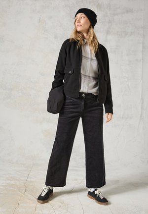 Giacca nera con cerniera e tasche frontali, indossata sopra una leggera camicia a quadri. Pantaloni a gamba larga in velluto a coste neri e sneakers nere con lacci bianchi.