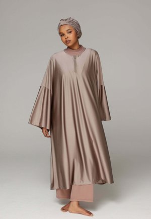 Femme portant une robe longue à manches longues en satin taupe et un turban assorti, debout pieds nus devant un fond gris uni.