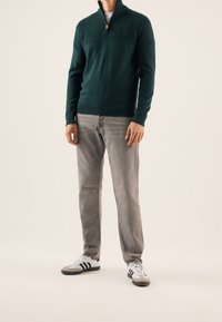 Donkergroene gebreide trui met een hoge kraag, voorzien van een ritssluiting en een gestructureerd ontwerp, gecombineerd met lichtgrijze jeans en witte sneakers.