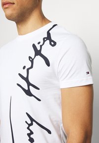 Camiseta de algodón blanca con un gran diseño gráfico negro en el frente y un pequeño acento de logo en la manga. Estilo clásico de cuello redondo.