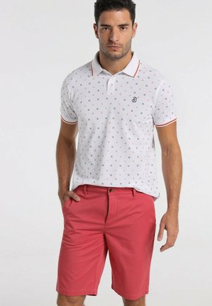 Polo shirt blanc avec de petits motifs géométriques multicolores, accents rouges sur le col ; associé à un short rouge de longueur genou.