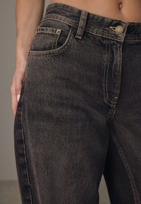 Jeans de mezclilla negra con una textura desgastada, con costuras amarillas, un bolsillo frontal y un cierre de botón en la cintura.