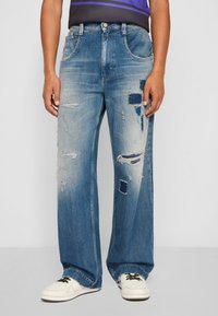 Light blue denim jeans with a relaxed fit, featuring distressed patches, frayed details, and wide legs. Paired with white sneakers.