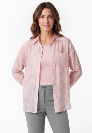 Frau mit schulterlangen braunen Haaren trägt ein hellrosa Hemd mit Knöpfen über einem pinken Top und grauen Hosen und steht mit den Händen hinter dem Rücken.