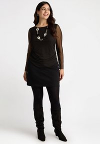 Blusa negra con mangas largas transparentes, diseño en capas, falda negra y medias negras. Accesorios incluyen un collar plateado grueso y botas altas hasta la rodilla.