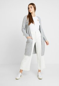 Grauer Cardigan aus weichem Stoff, offen designt, mit langen Ärmeln und seitlichen Taschen, kombiniert mit weißen weiten Hosen und weißen Sneakers.