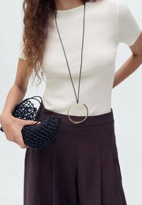Zwarte geweven handtas in de linkerhand; gouden ronde hanger ketting; aansluitend crèmekleurig top; donkere geplooide broek; lang, krullend haar.