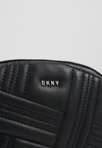 Accessoire en cuir noir texturé avec surpiqûres matelassées et petites lettres argentées de la marque DKNY centré près du bord supérieur.