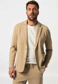 Blazer beige con una texture morbida, due tasche frontali e chiusura a un bottone, abbinato a una t-shirt bianca e pantaloni coordinati.