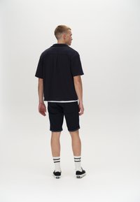 Schwarzes, kurzärmliges Hemd mit Kragen, kombiniert mit dunklen Shorts, weißen Socken und schwarzen Schuhen. Einfaches, lässiges Design ohne Muster.