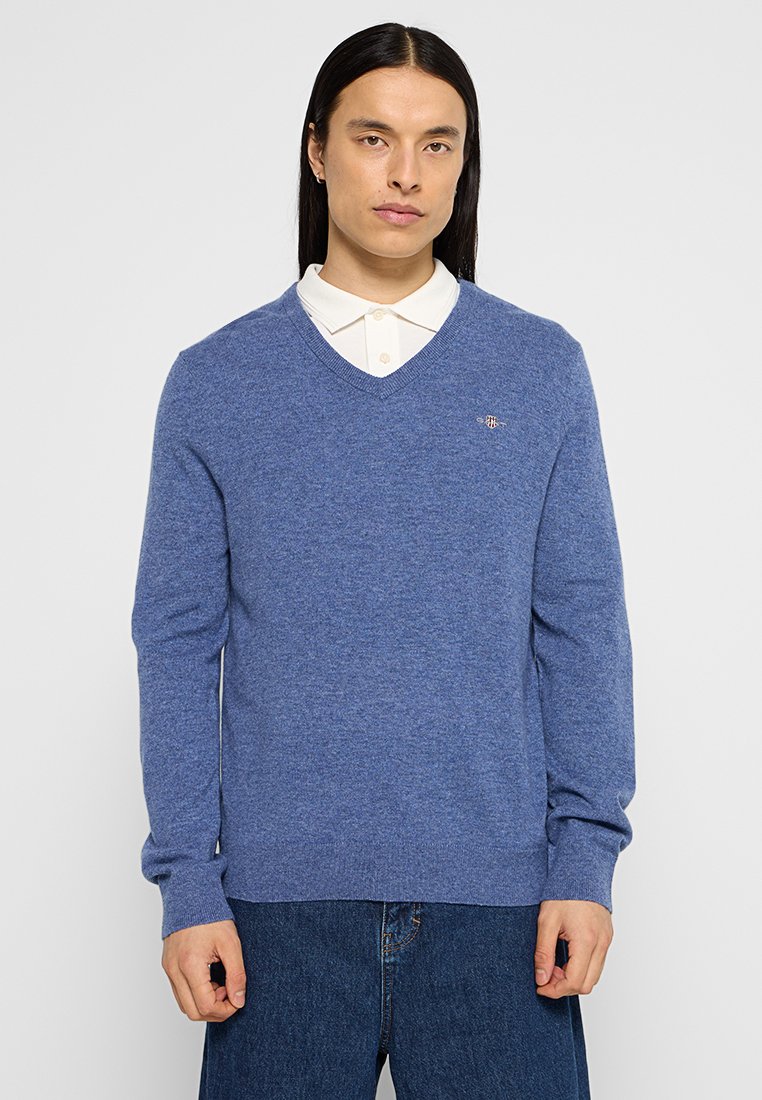 Niebieski sweter w serek z dzianiny, z długimi rękawami i małym logo na piersi. Nosi się go na białej koszuli z kołnierzykiem.