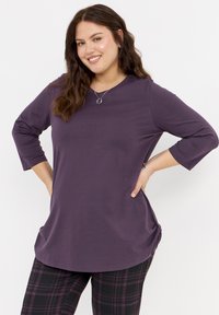 Lila Langarm-Oberteil mit rundem Ausschnitt und seitlichem Raffen, kombiniert mit schwarz-lila karierten Leggings. Weicher Stoff, lockere Passform.