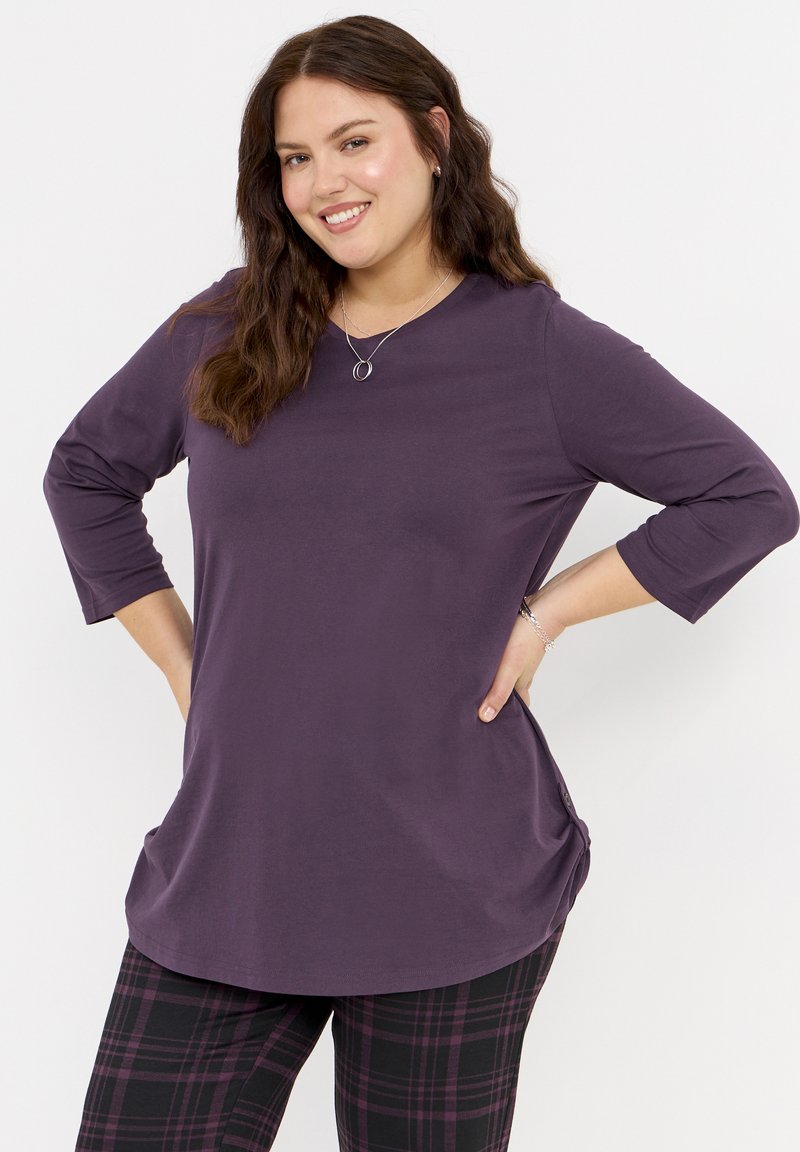 Lila Langarm-Oberteil mit rundem Ausschnitt und seitlichem Raffen, kombiniert mit schwarz-lila karierten Leggings. Weicher Stoff, lockere Passform.