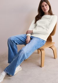 Bílý žebrovaný svetr, volný střih, v kombinaci se svetříkovými džíny světle modré barvy. Model sedí na dřevěné židli a má na sobě bílé tenisky. Jednoduchý, neformální outfit.