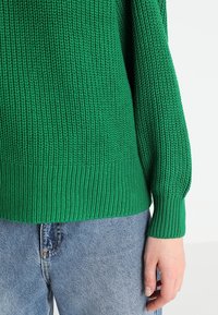 Maglione verde lavorato a maglia con un motivo a coste testurizzate e maniche lunghe, abbinato a jeans in denim azzurro chiaro. Polsi aderenti ai polsi.