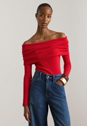 Červený top so zloženým výstrihom mimo plecia, dlhými rukávmi a priliehavým strihom, kombinovaný s modrými džínsami s vysokým pásom. Dopĺňa ho robustný náramok.