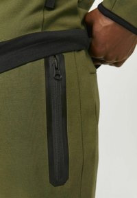 Olive green athletic pants feature a slim fit, black zippered pocket, and textured fabric with a smooth waistband for comfort.