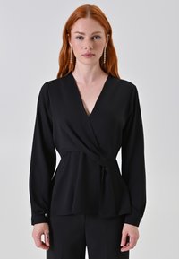 Blusa nera avvolgente realizzata in tessuto morbido, con maniche lunghe, scollo a V profondo e un dettaglio di vita legato per un silhouette fluida.