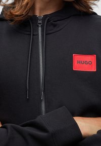 Černá mikina s kapucí na zip po celé délce, žebrované manžety a červený obdélníkový nášivka s nápisem "HUGO" v oblasti levého hrudníku. Hladký povrch.