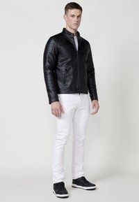 Giacca di pelle nera con dettagli testurizzati sulle spalle, chiusura con zip frontale e due tasche laterali, abbinata a jeans bianchi e sneakers nere.