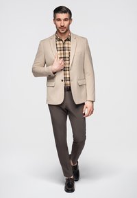 Homme portant un blazer beige, une chemise à carreaux, un pantalon foncé et des chaussures noires, debout devant un fond clair, faisant face vers l'avant.