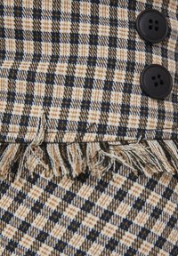 Tkanina tkana w kratkę, w odcieniach beżu, granatu i brązu. Posiada dwa czarne guziki i frędzle na krawędzi.