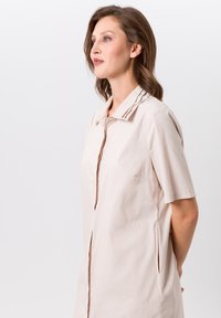 Robe chemise beige clair à manches courtes avec col pointu et fermeture à boutons sur le devant. Elle possède des poches latérales et des accents de couture subtils.