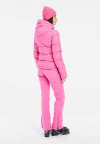 Giacca piumino rosa con cappuccio, design fitted. Pantaloni rosa coordinati con cerniere alle caviglie. Stivali da sci neri con dettagli di branding.