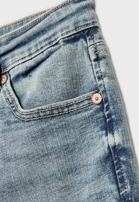Jeans di denim azzurro chiaro con un primo piano di una tasca caratterizzata da cuciture a contrasto e un bottone metallico decorativo. La texture appare usurata e sbiadita.