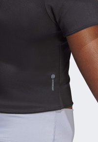 Kortärmad, svart atletisk tröja tillverkad av mjukt tyg. Har en slim fit med "AEROREADY"-logotyp nära fållen och en rundad nederkant.