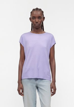 Kvinde med fletninger, der bærer en kortærmet lavendel top og lyseblå jeans, stående mod en ensfarvet hvid baggrund.