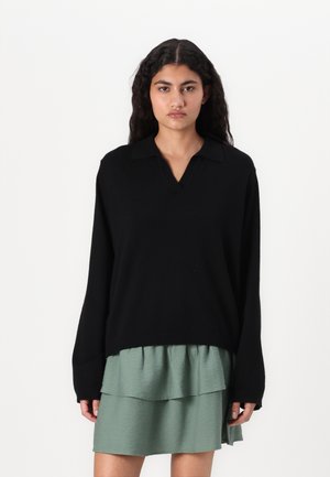Maglione nero oversize con scollo a V e maniche lunghe indossato sopra un vestito leggero verde chiaro a strati, caratterizzato da un tessuto testurizzato.
