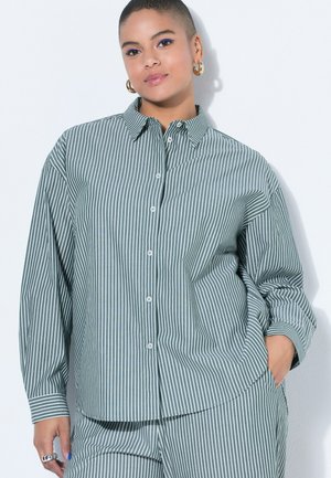 Osoba z krótkimi włosami, ubrana w zielono-białą koszulę zapinaną na guziki w pionowe paski oraz pasujące spodnie, pozująca z ręką w kieszeni.